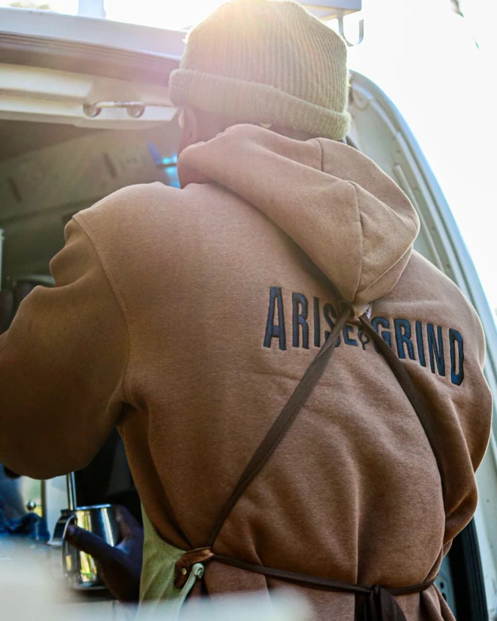 The embroidered Arise & Grind wordmark on a barista's brown hoodie, lit by morning sun beside the coffee van.
