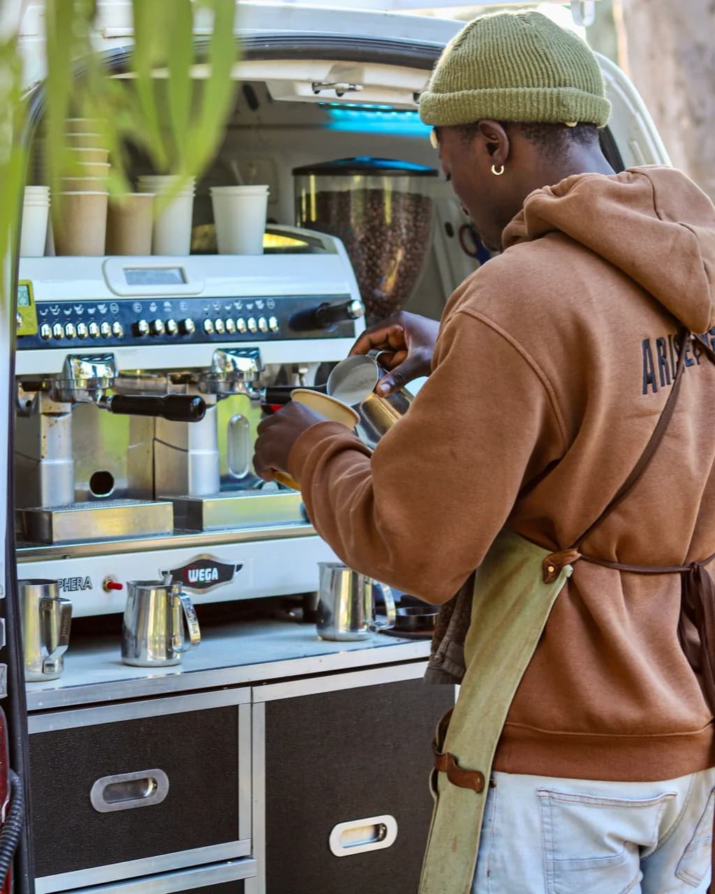 An Arise & Grind barista in a green beanie pours milk into a cup beside the espresso machine in the coffee van.